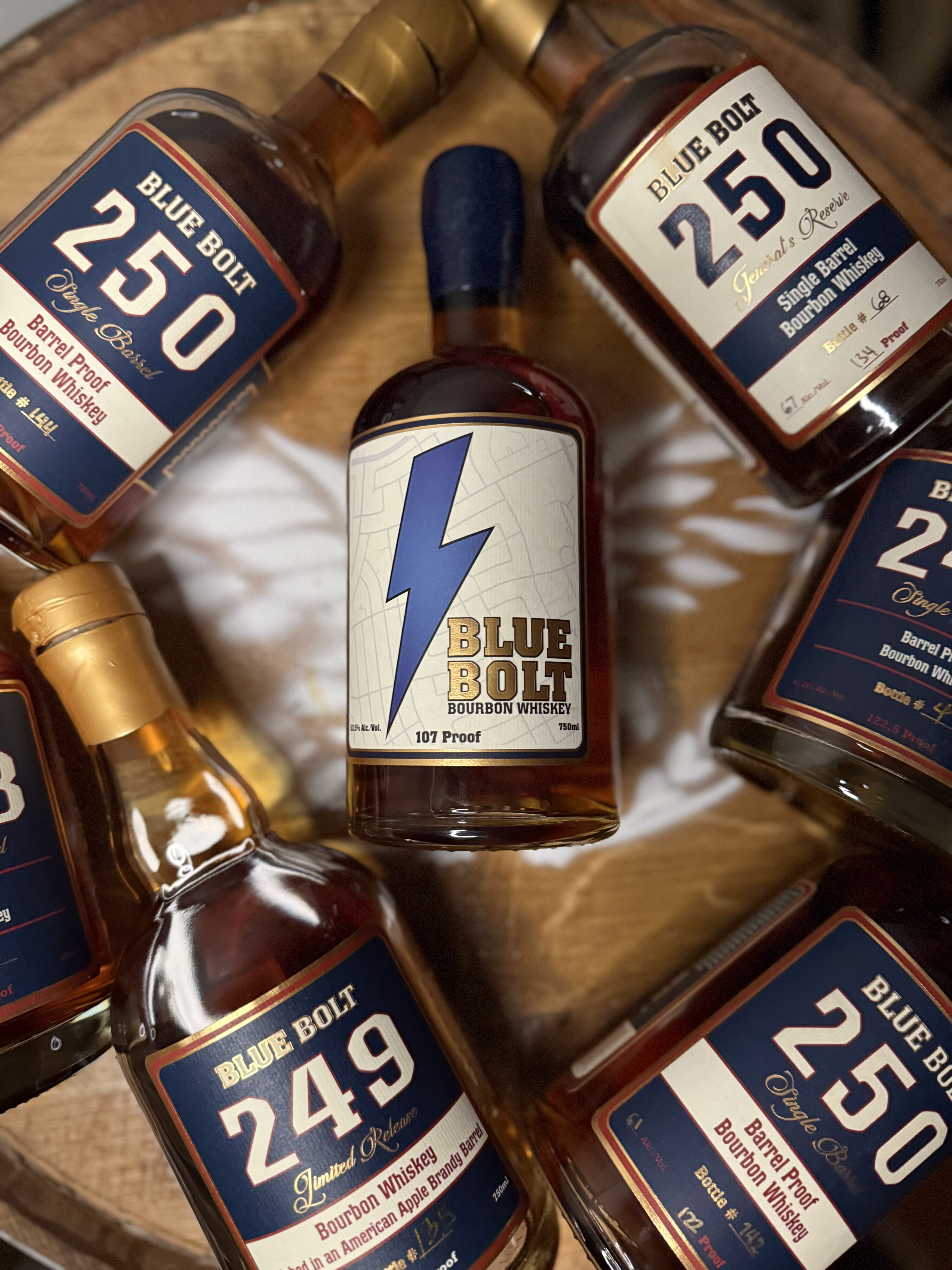 Blue Bolt 107 Proof Small Batch Bourbon lying on whiskey barrel with past single barrels surrounding it
