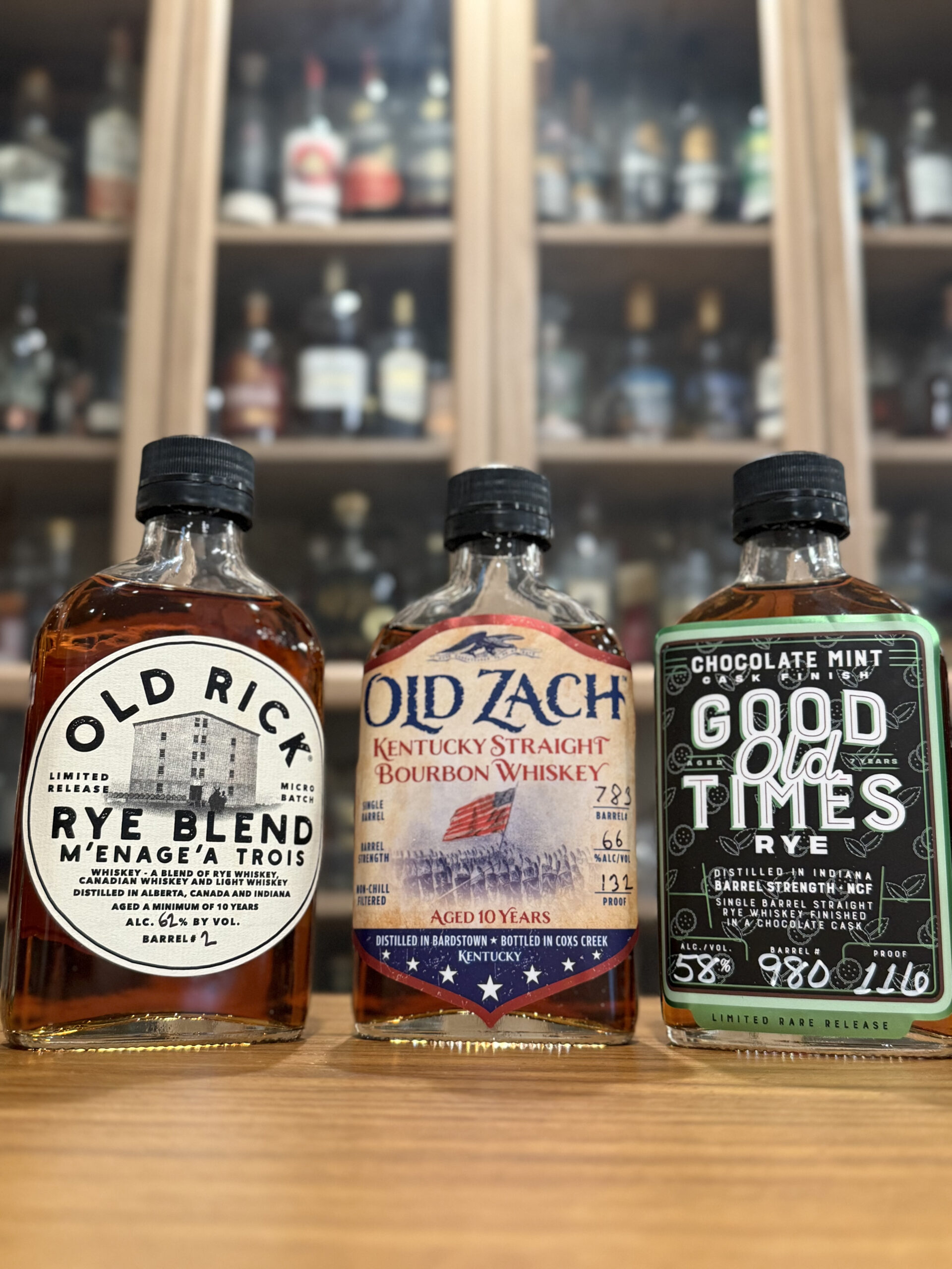 Three Cox's Creek Distilling Samples: Old Zach 10 Year Single Barrel Kentucky Bourbon 132 Proof, Old Rick Menage A Trois Rye Blend 124 Proof and Good Times Chocolate Mint Cask Finished Rye 116 Proof placed on desk in front of blurred whiskey collection.