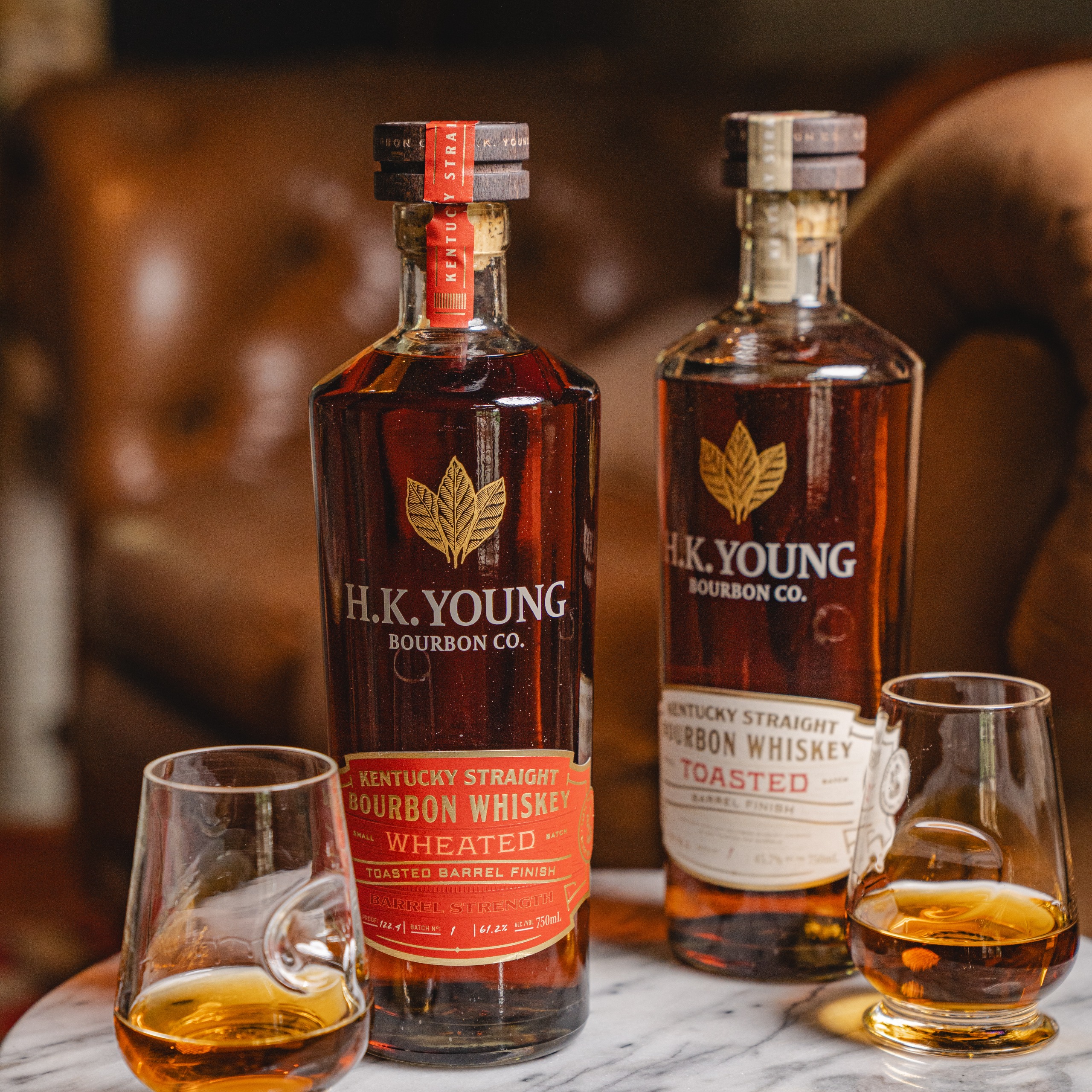 H.K. Young Toasted Wheated Bourbon Barrel Strength and 91.4 Proof Small Batch Toasted Bourbon on marble table with a full glass beside each bottle.