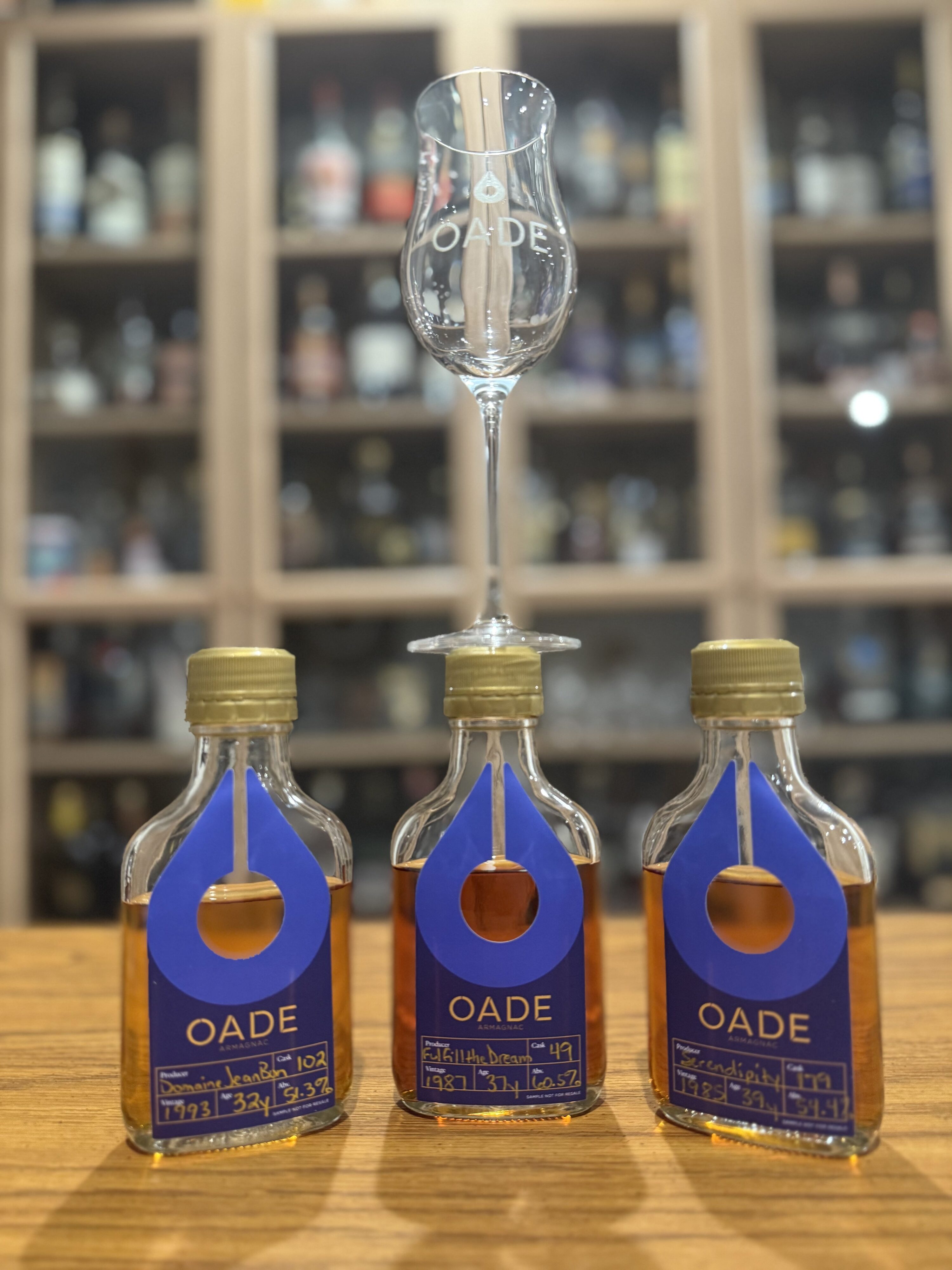 Three oade samples on desk with tasting glass balanced on top in front of liquor collection