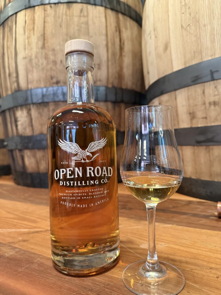 Open Road unlabeled bottle beside tasting glass in front of whiskey barrels