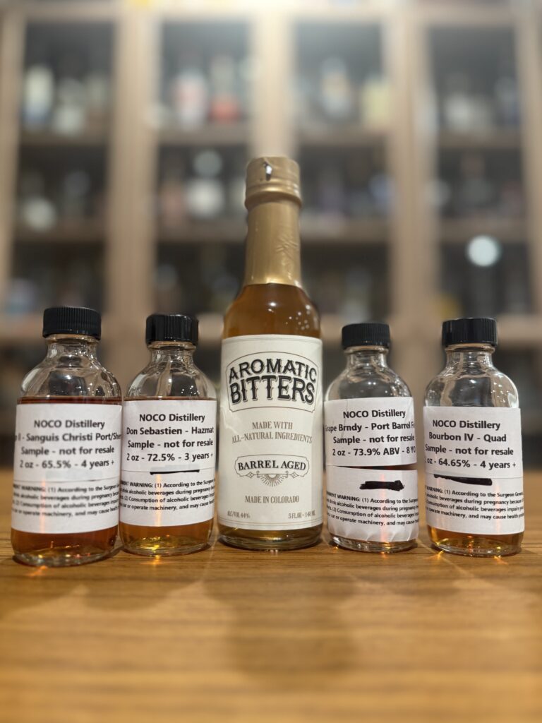 Noco Distillery four spirit samples and aromatic bitters on desk in front of whiskey collection