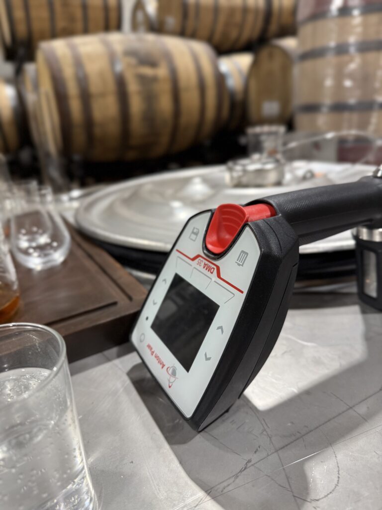 Hydrometer sits on metal storage tank in front of tasting glasses and whiskey barrels