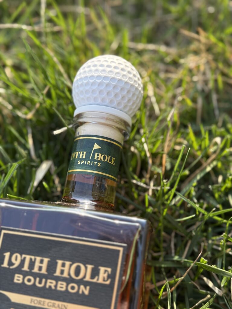 19th Hole Bourbon golf ball topper lying in grass