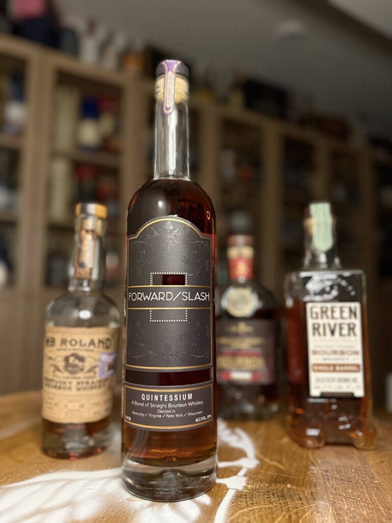 Forward Slash Quintessium standing on front of blurred sourced distillery bottles