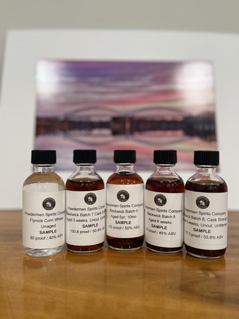 Powdermen Early Samples of Batches 6-9 and the Flyrock unaged whiskey with a picture of Arrigoni Bridge in Middletown, CT behind it.