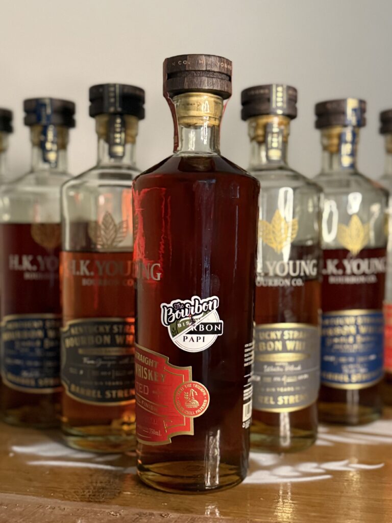 H.K. Young "The Bourbon Bishop x Bourbon Papi" Toasted Wheated Bourbon Single Barrel standing in front of descending rows of other H.K. Young releases