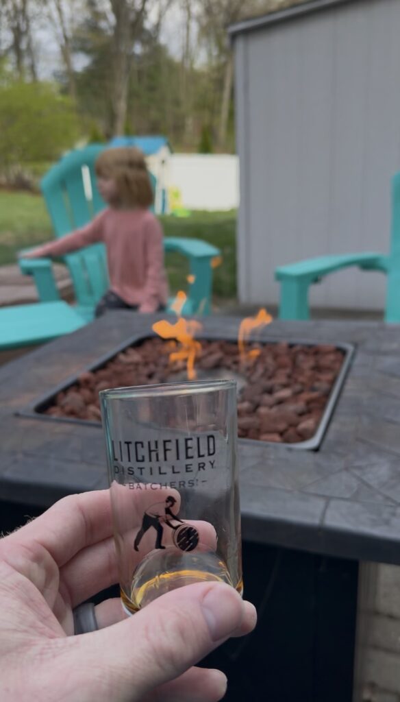 Litchfield distillery tasting glass with whiskey in front of fire and child playing in the yard