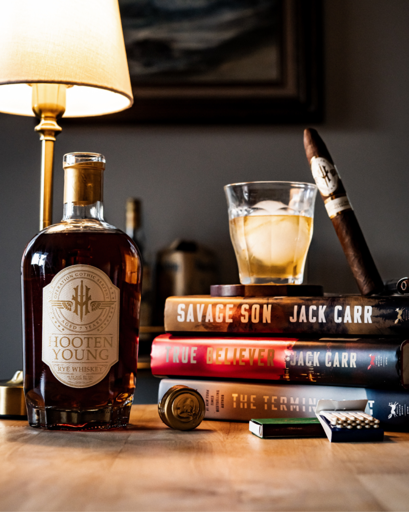 Hooten Young's Gothic Serpent Rye sits open with a freshly poured glass with one large ice cube and paired cigar against on top of Jack Carr books