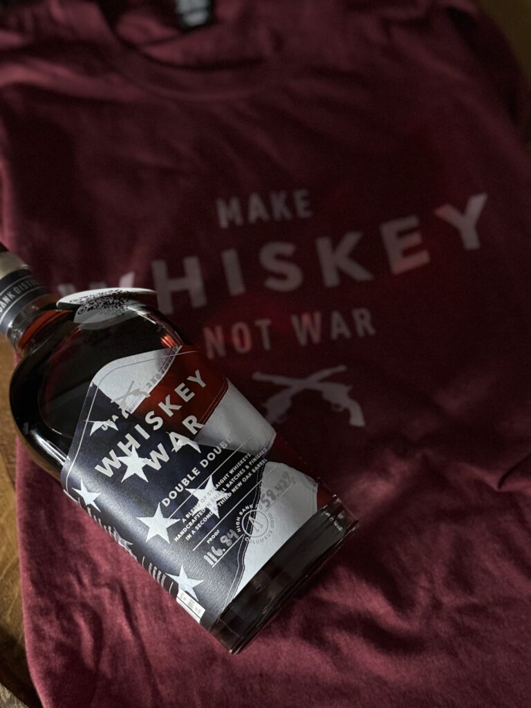 High Banks Whiskey War Double Double Oak 250th Anniversary Release lying on darkened "make whiskey not war" shirt