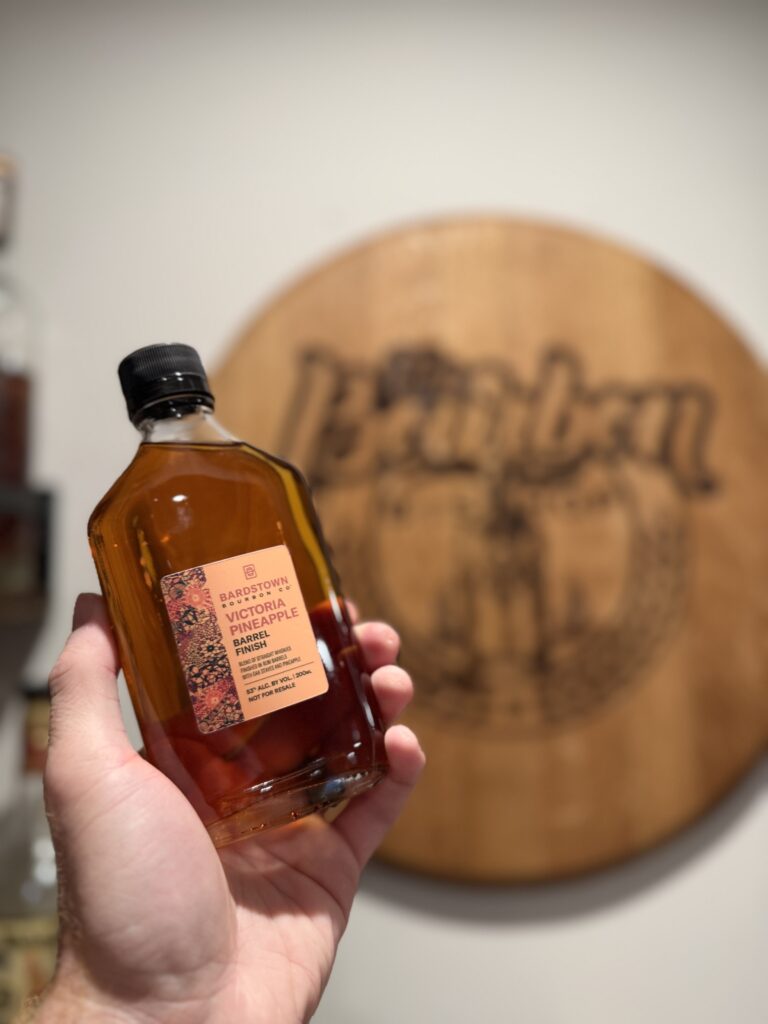 Bardstown Bourbon Company Victoria Pineapple sample held in front of the Bourbon Bishop barrel head made by Peter Staulis of Rekindled Barrels