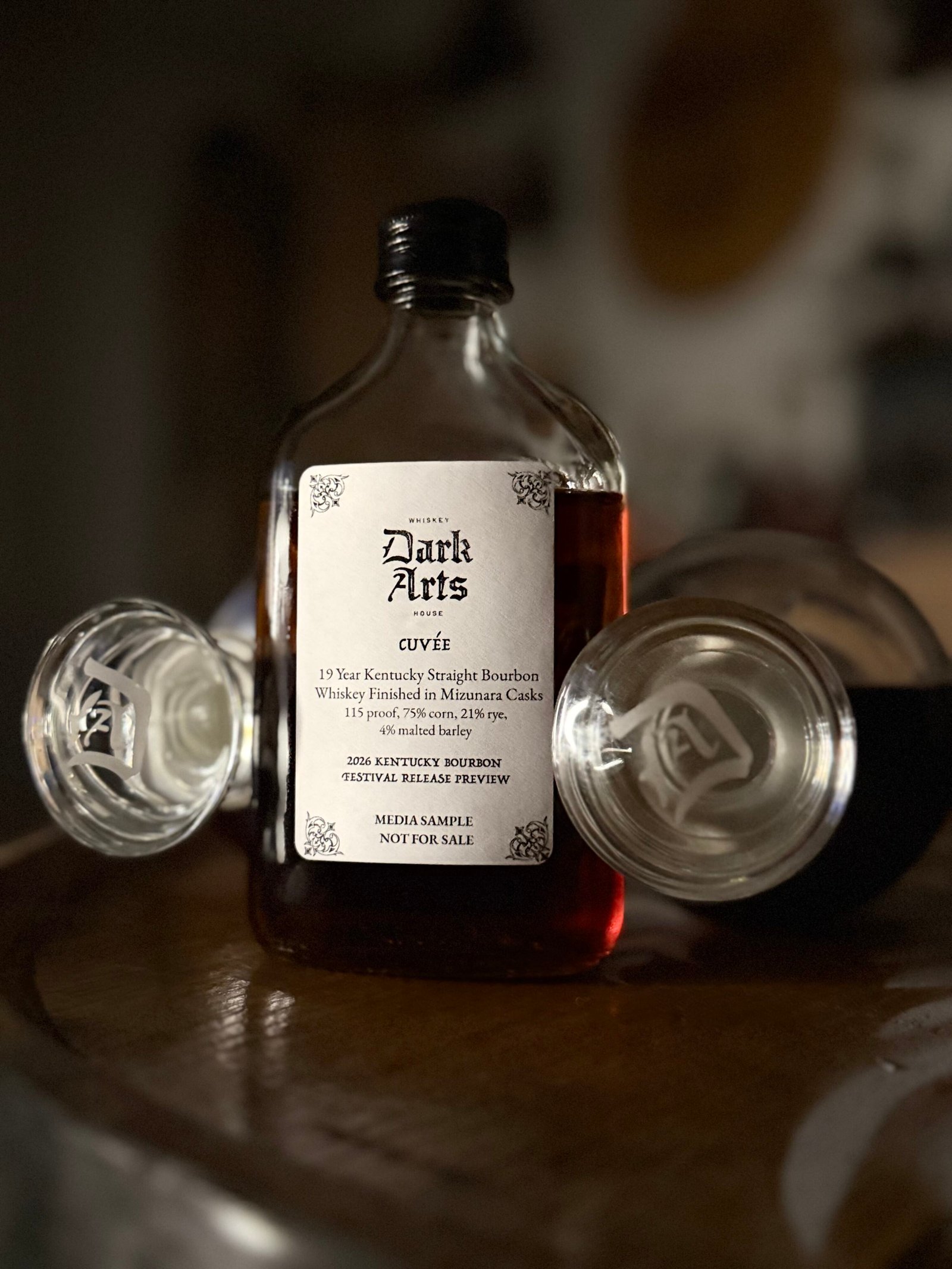 Dark Arts KBF Special Release: Chief Alchemist Reserve Cuvée 19 Year Kentucky Bourbon finished in Mizunara Casks stands between two Dark Arts bottle toppers