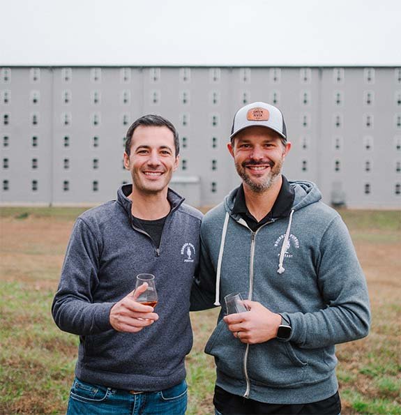 Founders of Bourbon Pursuit podcast and Pursuit Spirits Kenny Coleman & Ryan Cecil post in front of rickhouse.