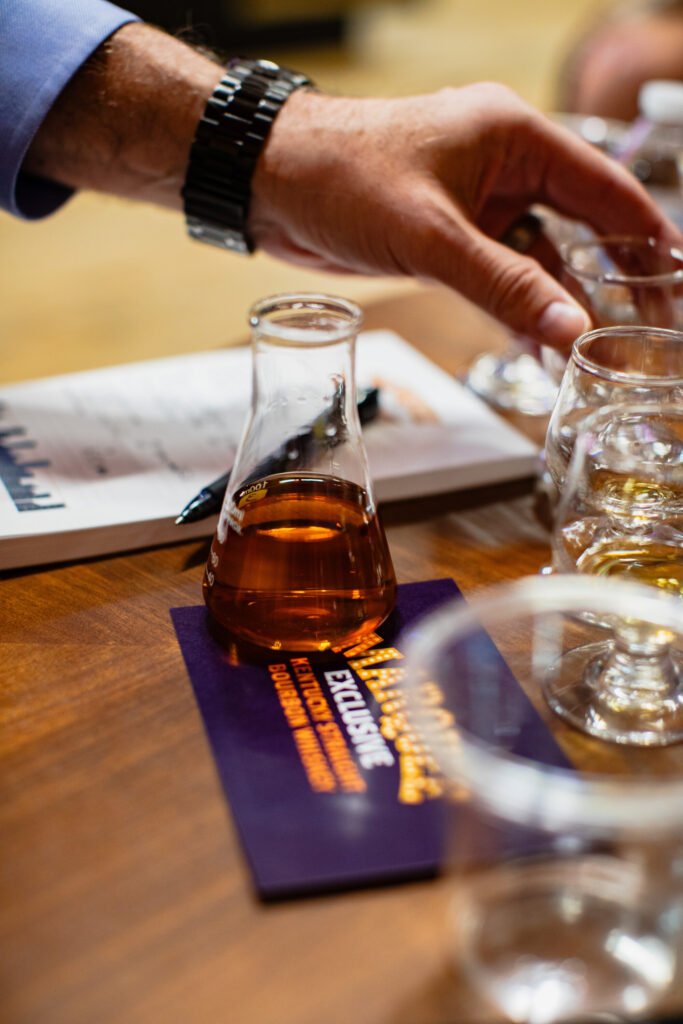 Hand reaches for tasting glasses by full beaker of bourbon on top of Marquee branded coaster