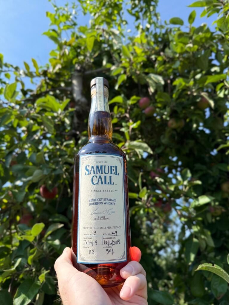 Hand holds Samuel Call Barrel 3 in front of apple tree full of ripe red apples