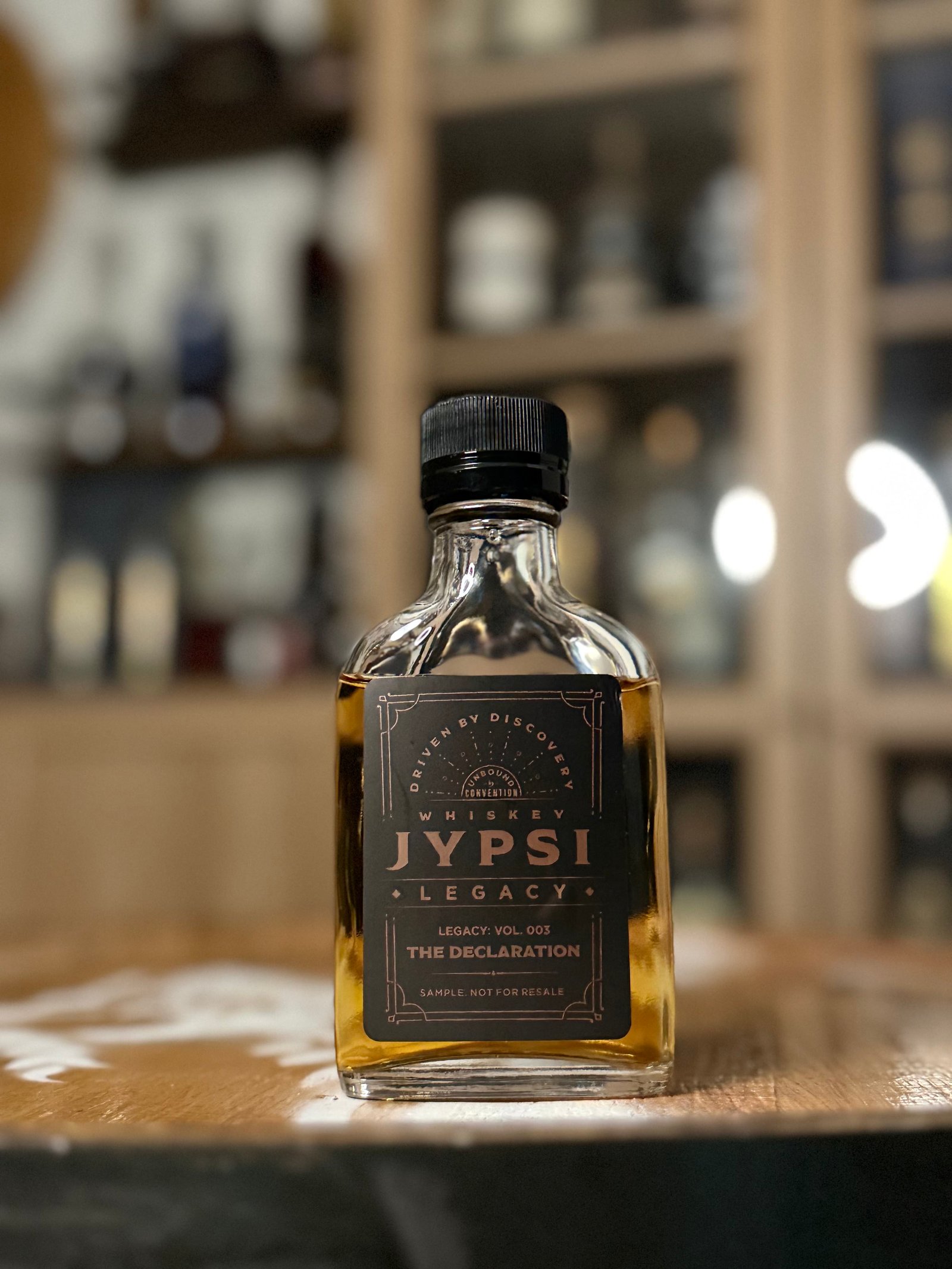 Whiskey JYPSI Legacy Batch 003 "The Declaration" stands in front of whiskey collection